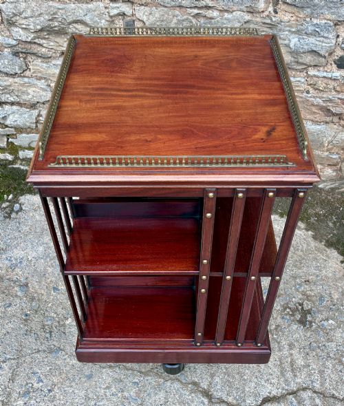 fig 1. An Edwardian mahogany revolving bookcase featuring a square top with a brass gallery rail, slatted sides with brass studs, two tiers of shelving, and a central rotating mechanism on castors.
