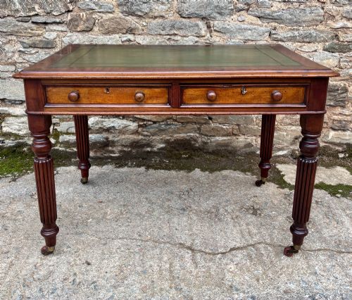 fig 1. A Victorian mahogany writing table featuring a green leather top, two front drawers with turned wooden knobs, and fluted turned legs on brass castors.