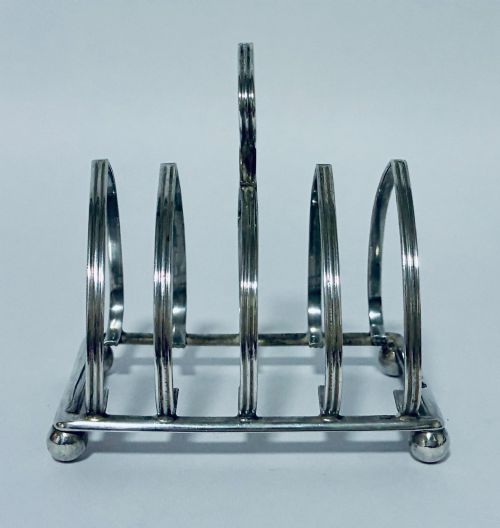fig 1. An antique silver toast rack featuring five arched divisions, a central loop handle, and a rectangular base supported by four ball feet.