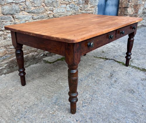 fig 2. A Victorian pine farmhouse table featuring a light natural timber plank top, two frieze drawers with round wooden knobs, and four dark turned wooden legs.