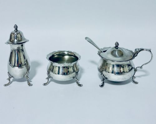 fig 1. A three-piece silver cruet set comprising a pepper caster, open salt cellar, and mustard pot with a spoon, each resting on shell-adorned feet.