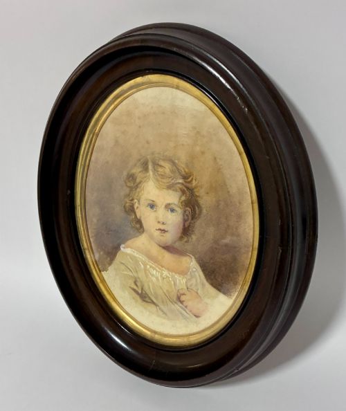 fig 1. An oval Edwardian watercolour portrait depicting a young child with blonde curls, set inside a dark wooden frame with a gilt inner trim.