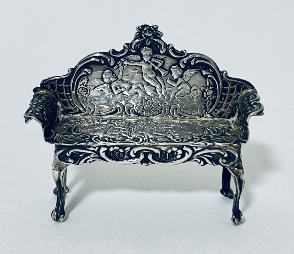 An ornate miniature sterling silver sofa featuring embossed cherub motifs, scrollwork backrest, and cabriole legs.