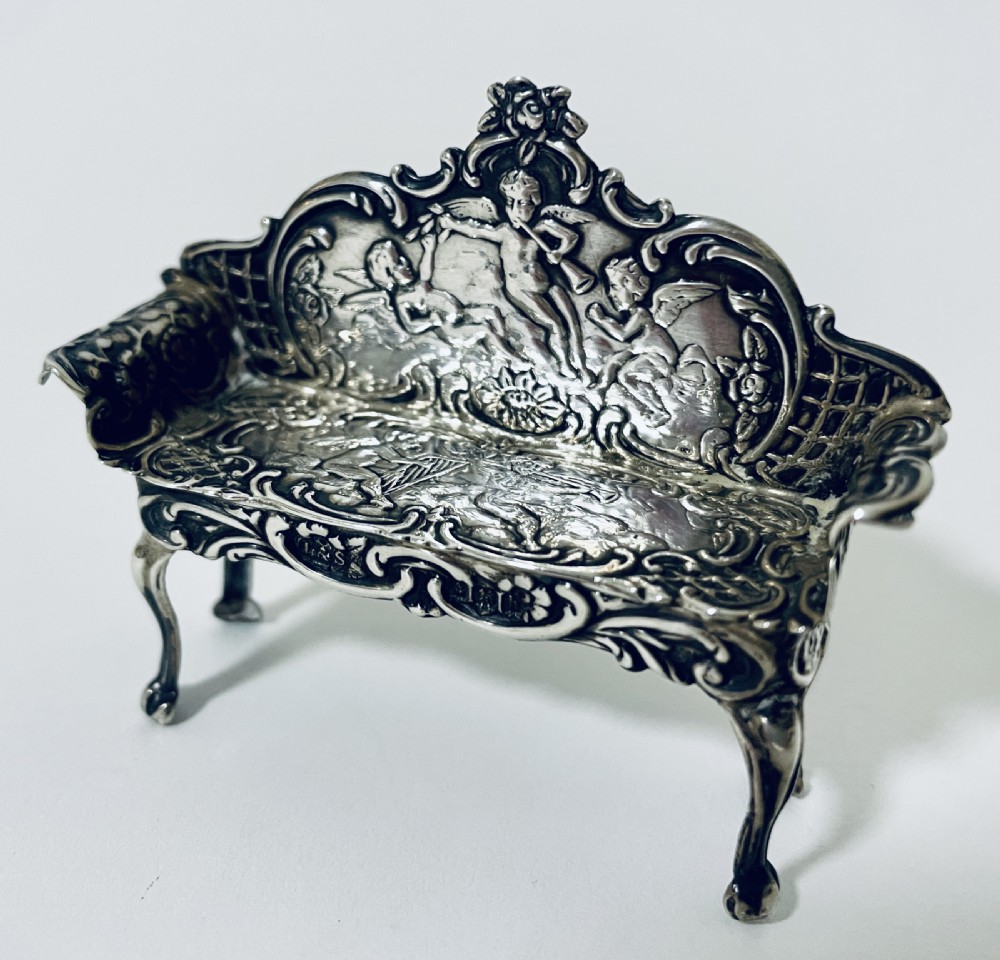 An ornate miniature sterling silver sofa featuring embossed cherub motifs, scrollwork backrest, and cabriole legs. View III