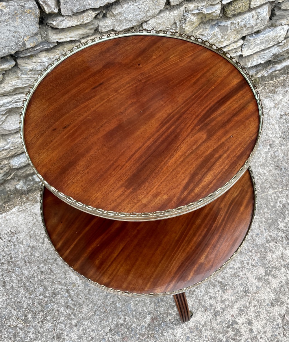 A Georgian dark mahogany two-tier dumb waiter featuring circular tops with pierced brass galleries, a central turned column, and sweeping reeded tripod legs on brass castors. View IX
