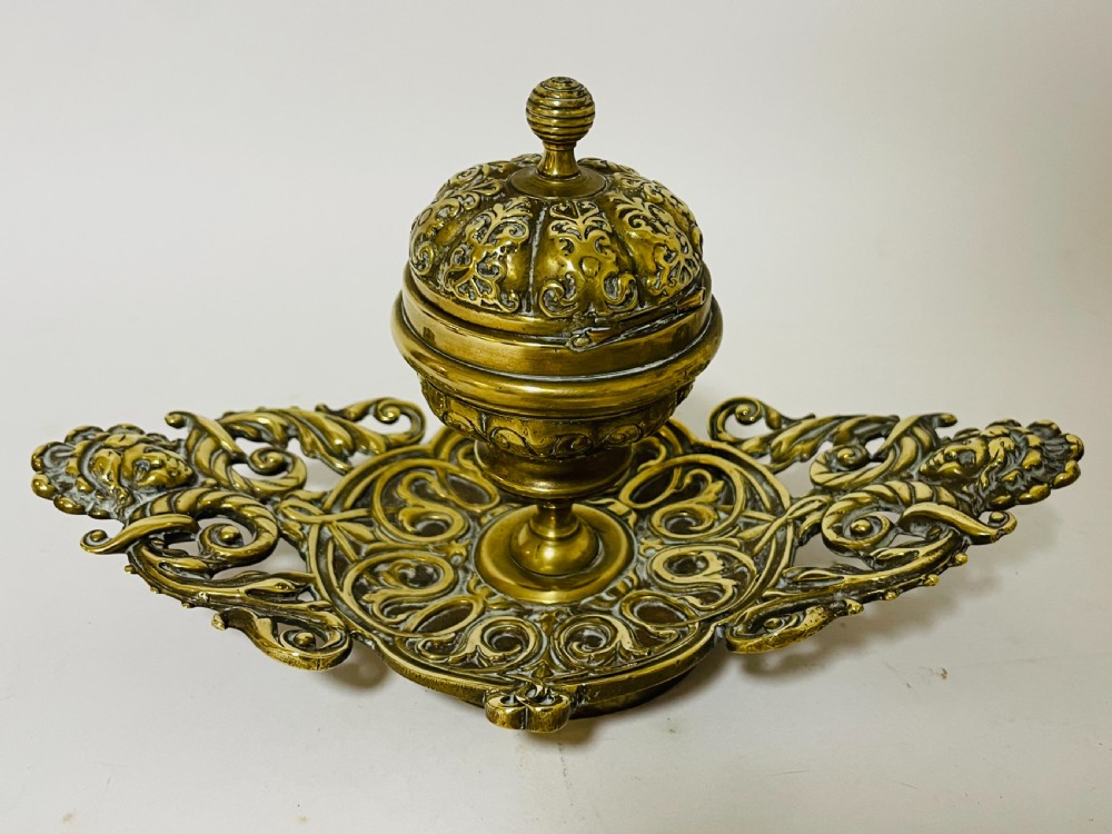 An ornate antique brass desk set featuring a lidded inkwell on a scrolled stand, a matching pen tray, and two turned brass candlesticks. View IX