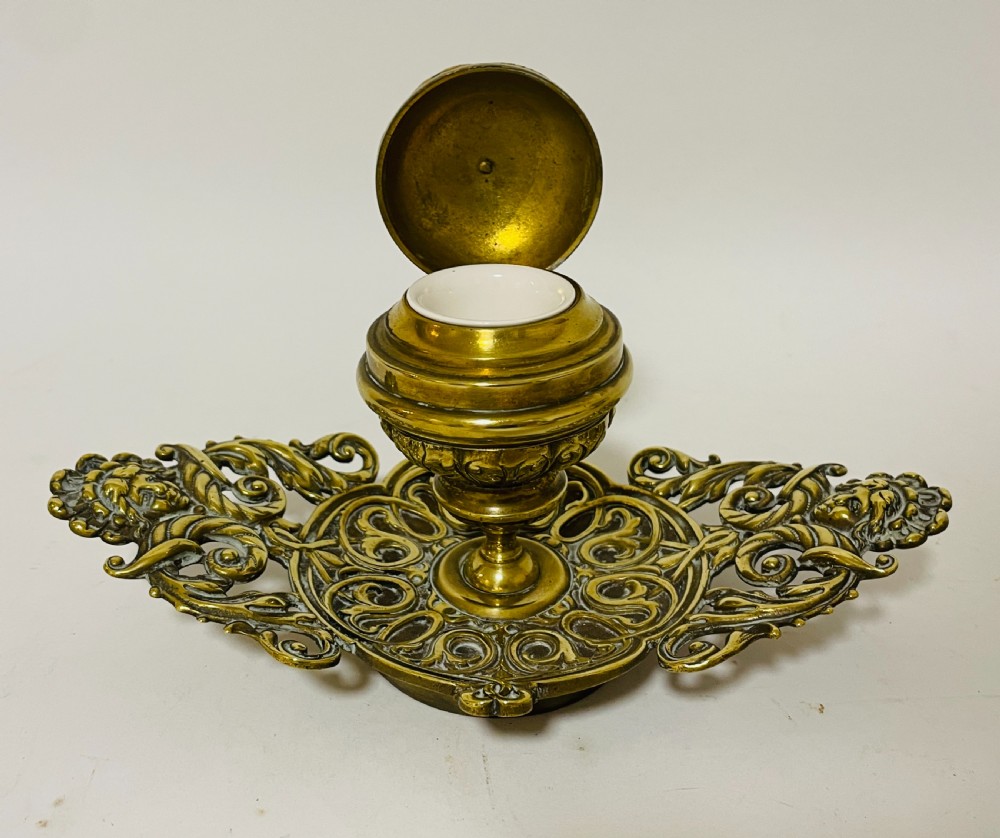 An ornate antique brass desk set featuring a lidded inkwell on a scrolled stand, a matching pen tray, and two turned brass candlesticks. View X