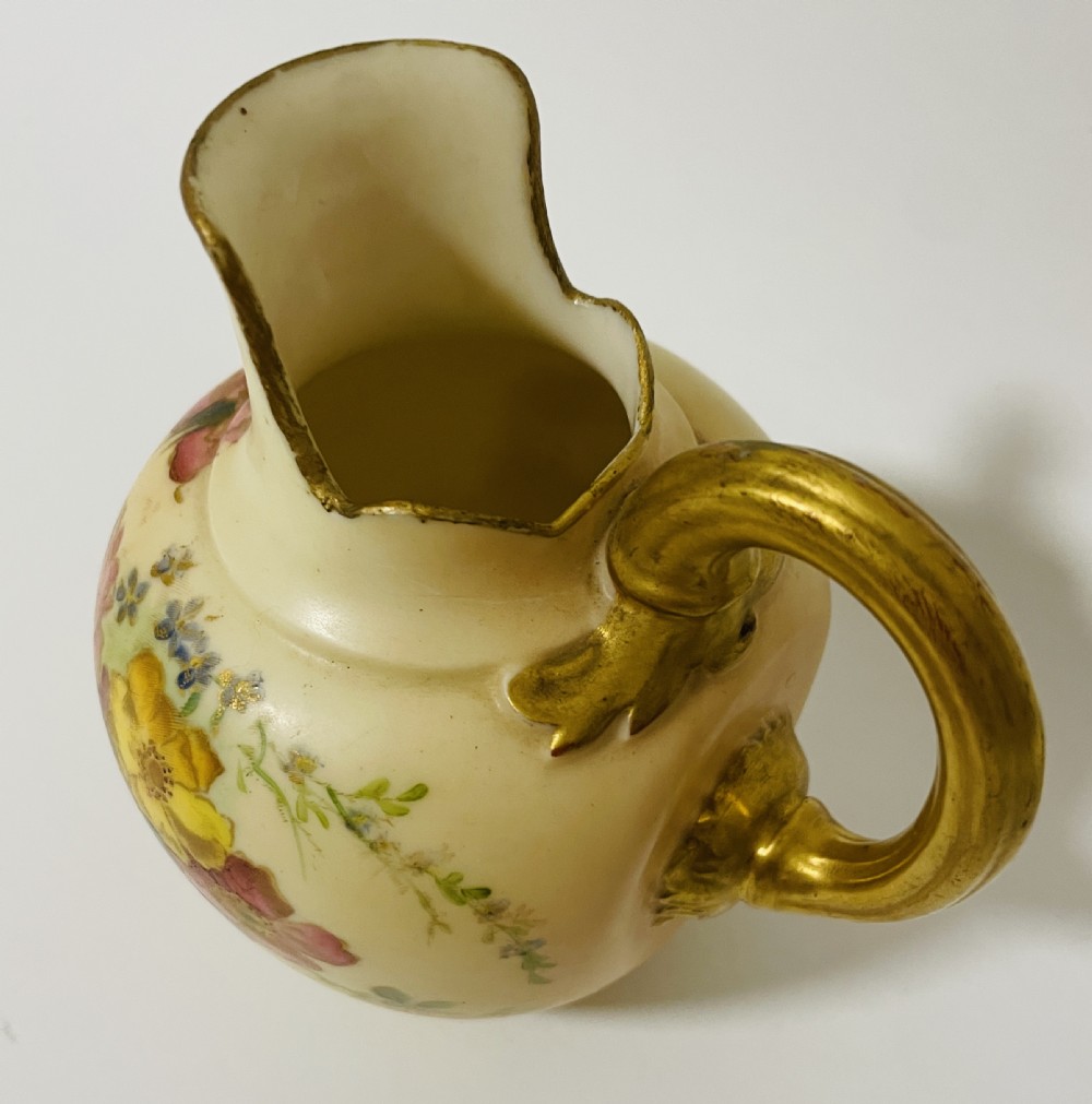 An Edwardian Royal Worcester porcelain jug featuring a blush cream body decorated with hand-painted yellow and pink flowers alongside a gilded handle and rim. View IX