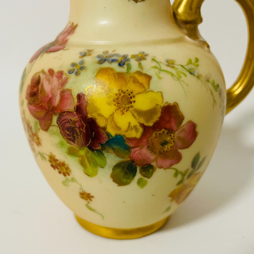 An Edwardian Royal Worcester porcelain jug featuring a blush cream body decorated with hand-painted yellow and pink flowers alongside a gilded handle and rim. View X
