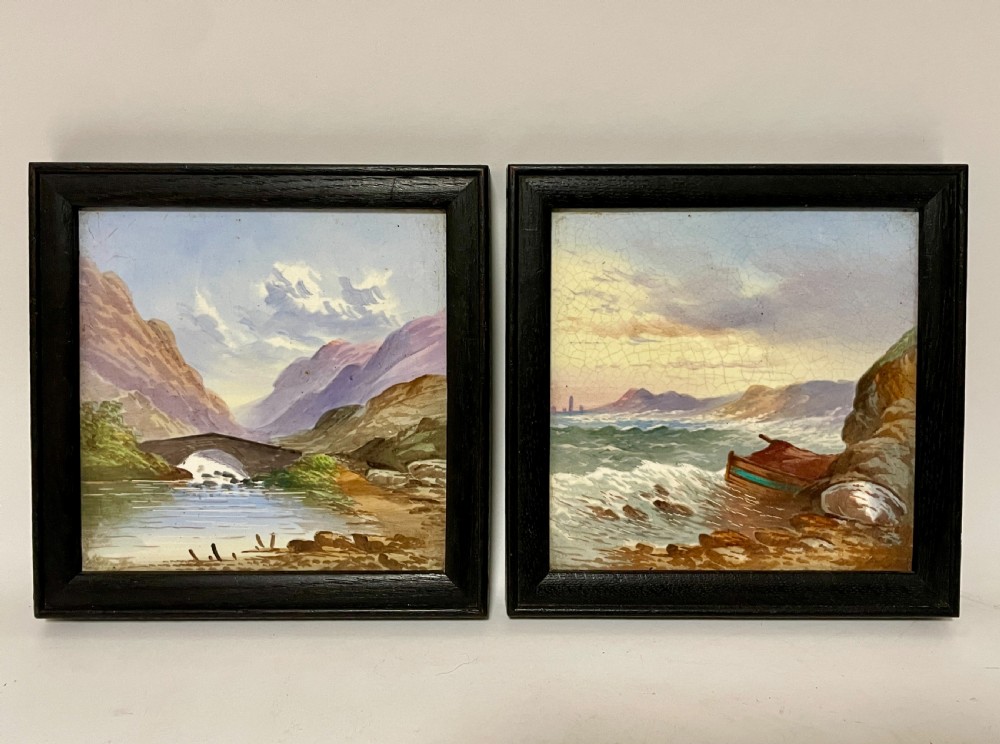 A pair of square Victorian ceramic tiles featuring hand-painted landscape scenes of a mountain river and a coastal shore, presented in dark wooden frames.