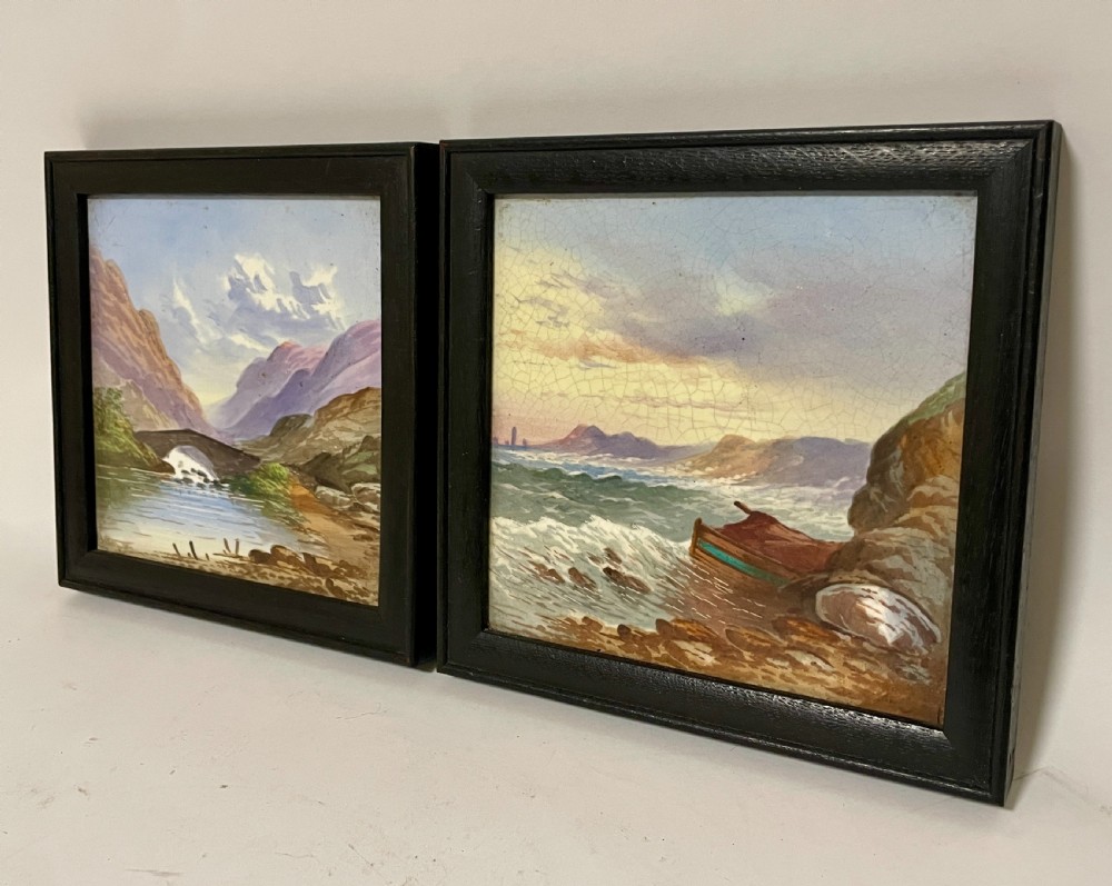A pair of square Victorian ceramic tiles featuring hand-painted landscape scenes of a mountain river and a coastal shore, presented in dark wooden frames. View II