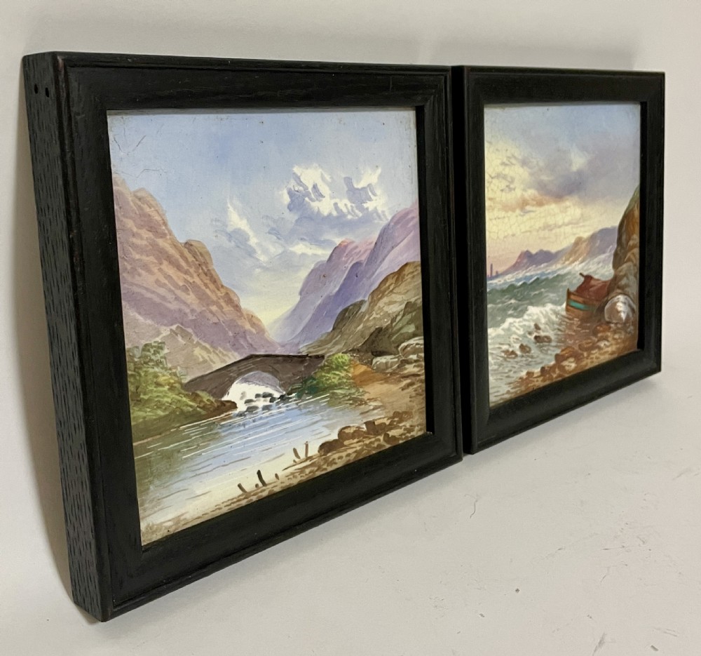 A pair of square Victorian ceramic tiles featuring hand-painted landscape scenes of a mountain river and a coastal shore, presented in dark wooden frames. View III