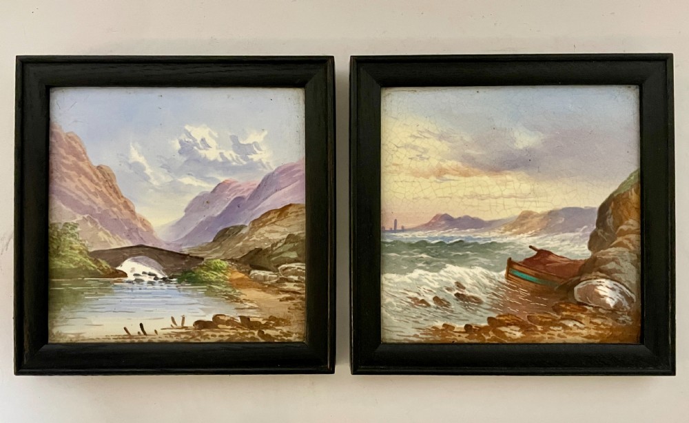 A pair of square Victorian ceramic tiles featuring hand-painted landscape scenes of a mountain river and a coastal shore, presented in dark wooden frames. View V