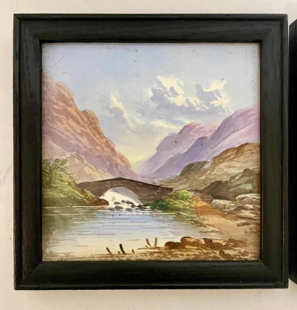 A pair of square Victorian ceramic tiles featuring hand-painted landscape scenes of a mountain river and a coastal shore, presented in dark wooden frames. View VI