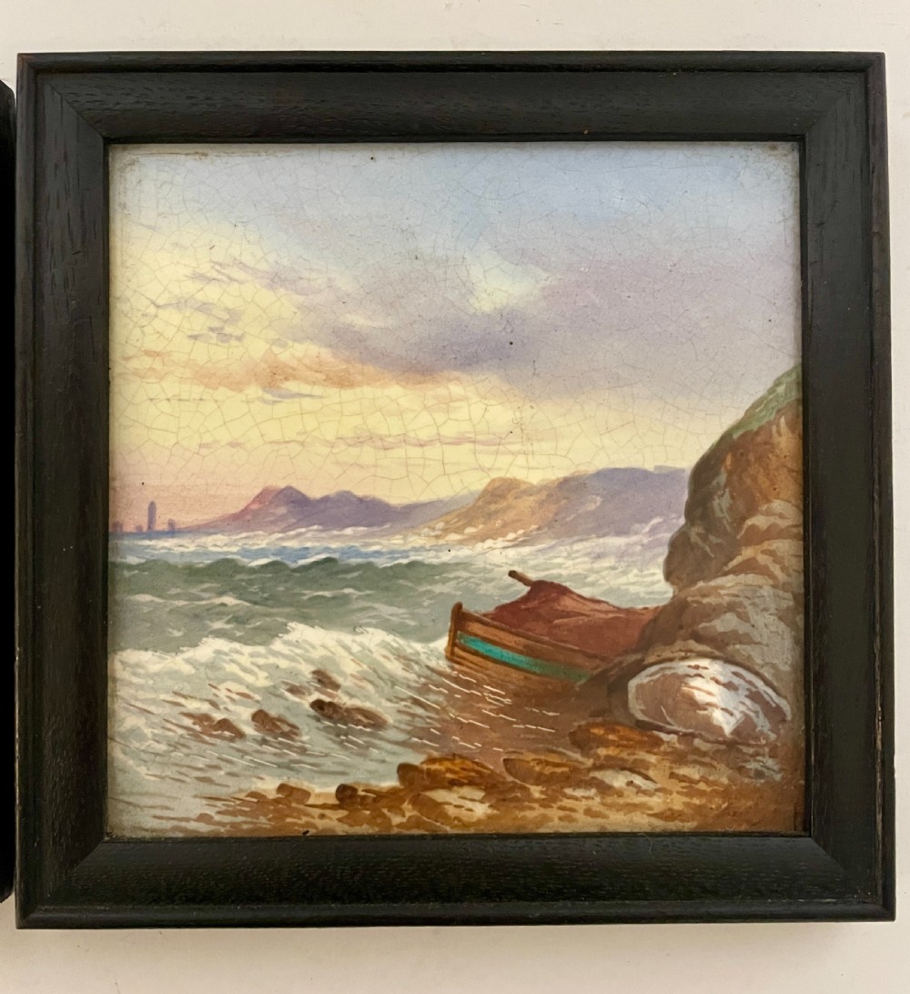 A pair of square Victorian ceramic tiles featuring hand-painted landscape scenes of a mountain river and a coastal shore, presented in dark wooden frames. View VII