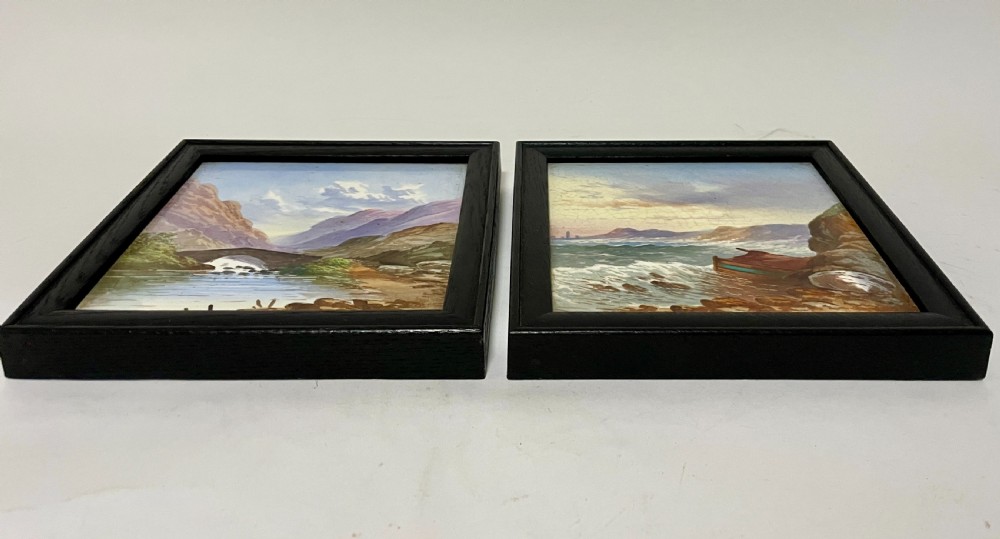 A pair of square Victorian ceramic tiles featuring hand-painted landscape scenes of a mountain river and a coastal shore, presented in dark wooden frames. View IX