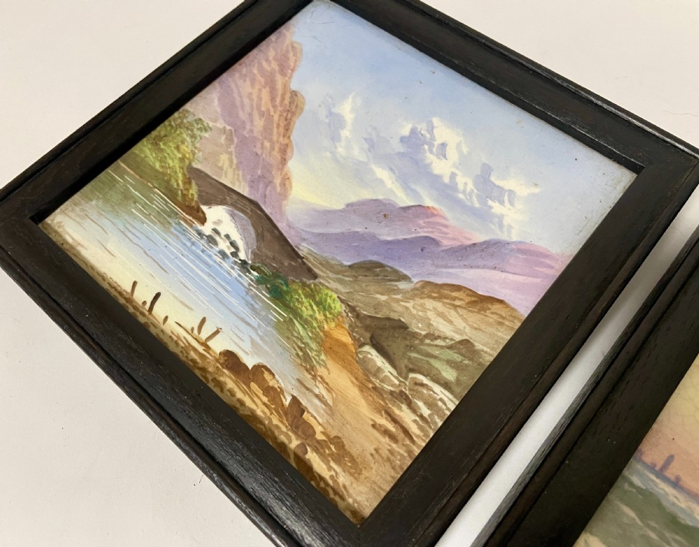 A pair of square Victorian ceramic tiles featuring hand-painted landscape scenes of a mountain river and a coastal shore, presented in dark wooden frames. View X