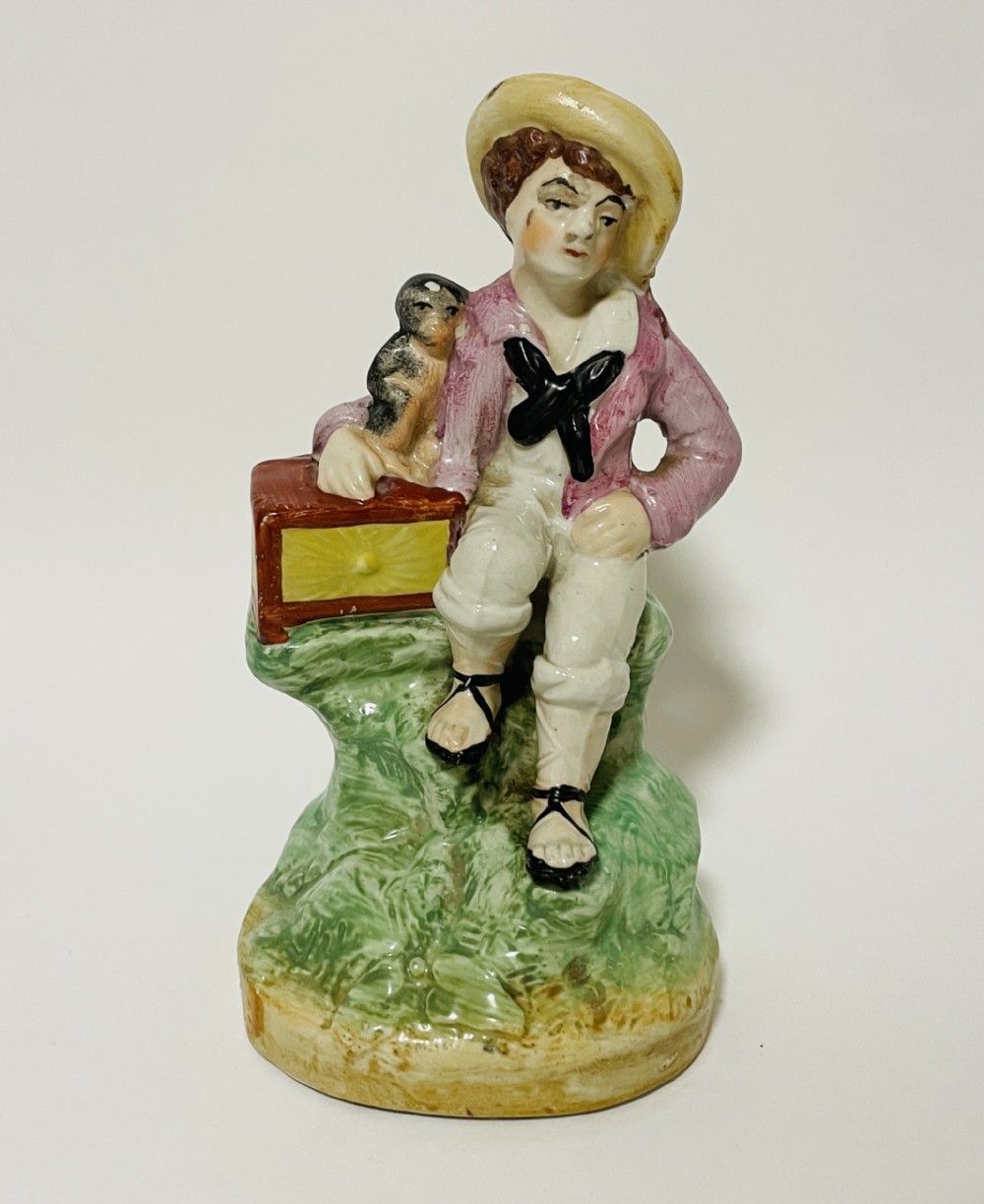 A Victorian Staffordshire earthenware figure depicting a seated boy in a straw hat and pink jacket alongside a monkey resting on a small brown chest.