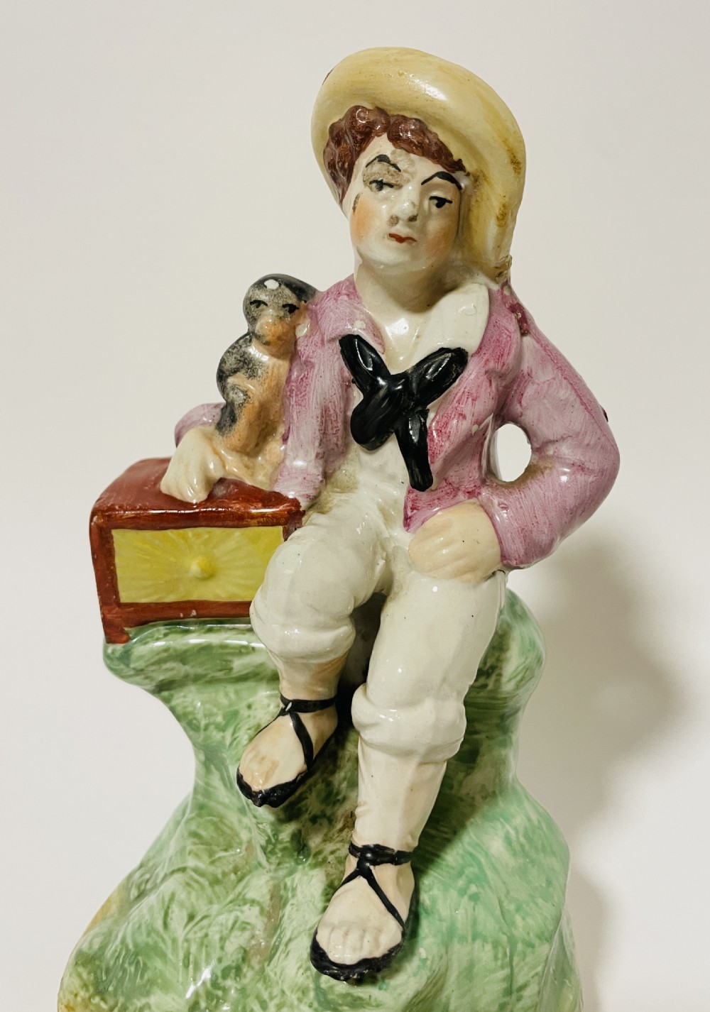 A Victorian Staffordshire earthenware figure depicting a seated boy in a straw hat and pink jacket alongside a monkey resting on a small brown chest. View III