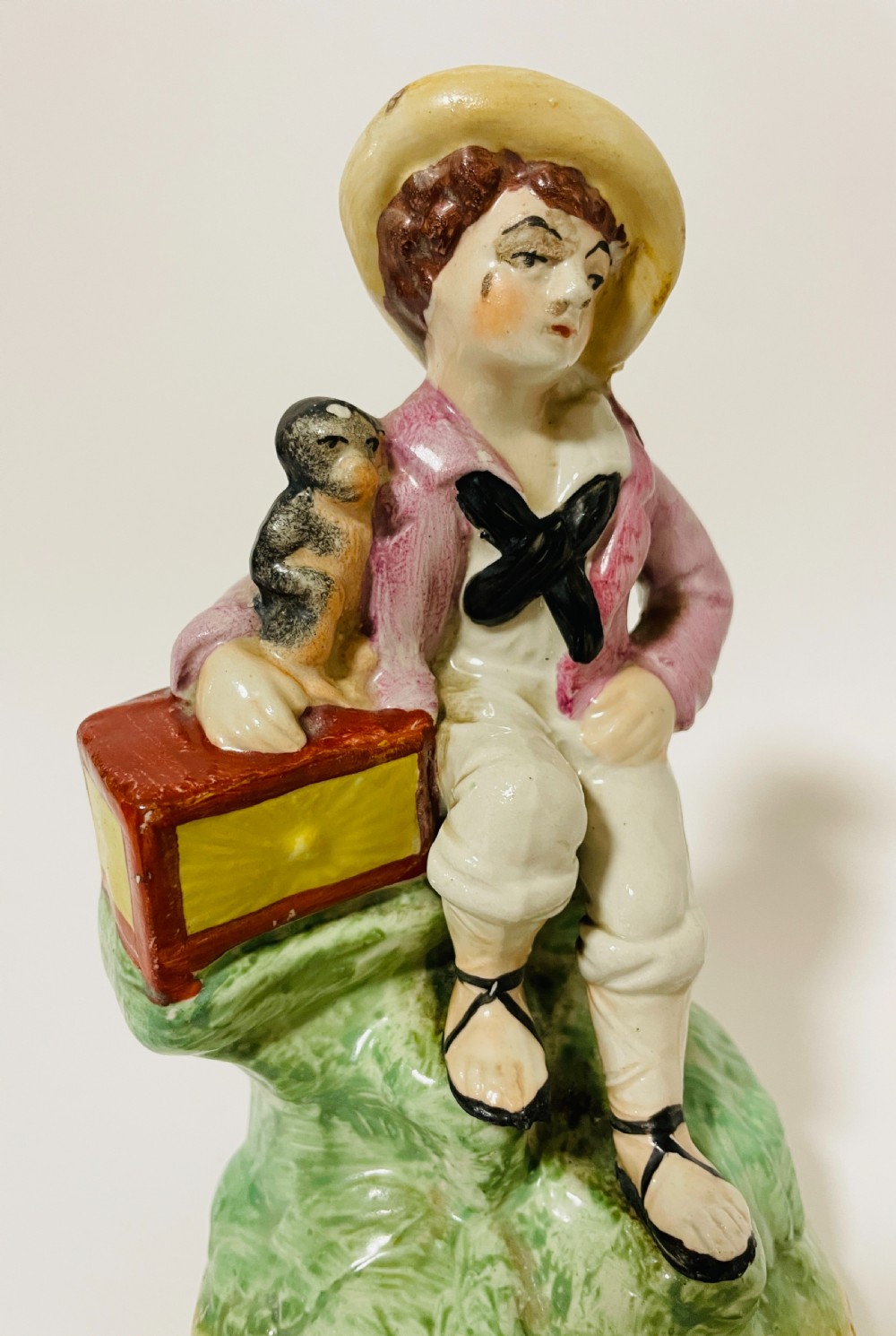 A Victorian Staffordshire earthenware figure depicting a seated boy in a straw hat and pink jacket alongside a monkey resting on a small brown chest. View X