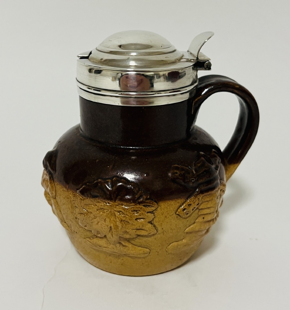 A Victorian two-tone brown stoneware mustard pot featuring relief-moulded rural scenes, a side handle, and a polished silver hinged lid.