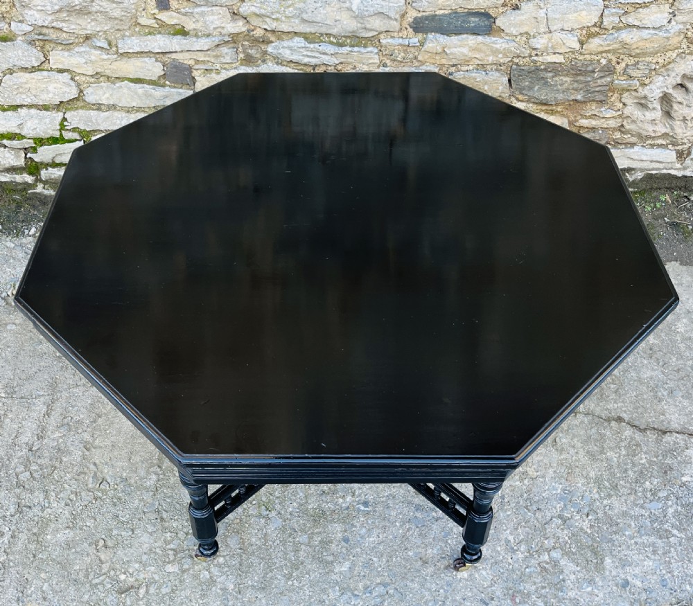 An octagonal ebonised Aesthetic Movement centre table featuring turned legs, brass castors, and a spindle X-frame stretcher. View II