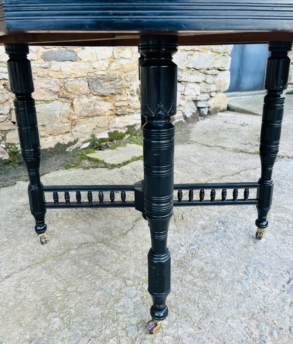 An octagonal ebonised Aesthetic Movement centre table featuring turned legs, brass castors, and a spindle X-frame stretcher. View V