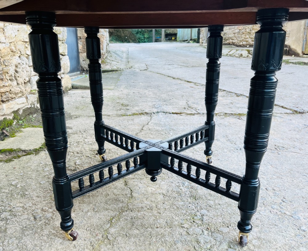 An octagonal ebonised Aesthetic Movement centre table featuring turned legs, brass castors, and a spindle X-frame stretcher. View VII