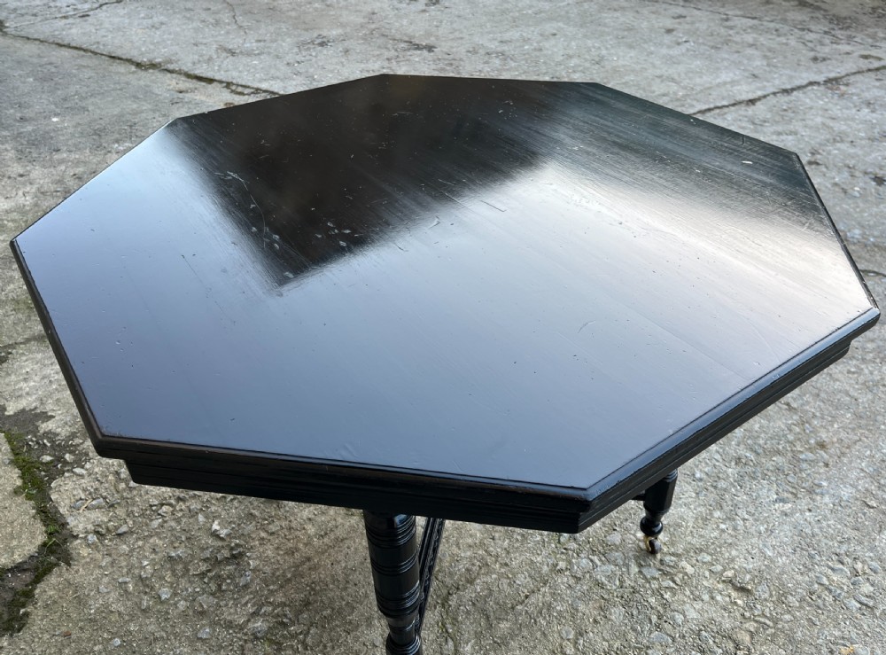 An octagonal ebonised Aesthetic Movement centre table featuring turned legs, brass castors, and a spindle X-frame stretcher. View XI