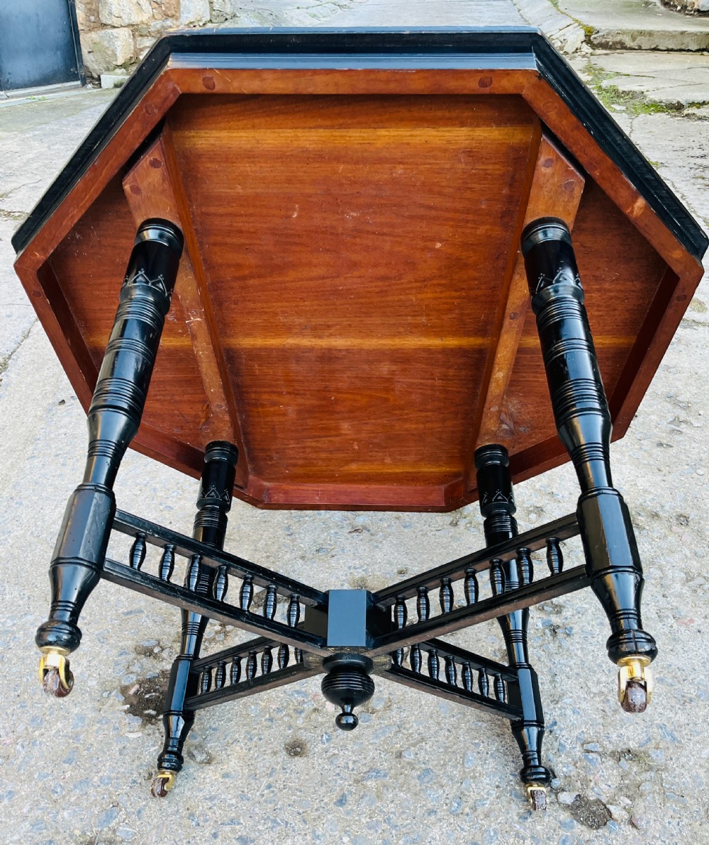 An octagonal ebonised Aesthetic Movement centre table featuring turned legs, brass castors, and a spindle X-frame stretcher. View XIII