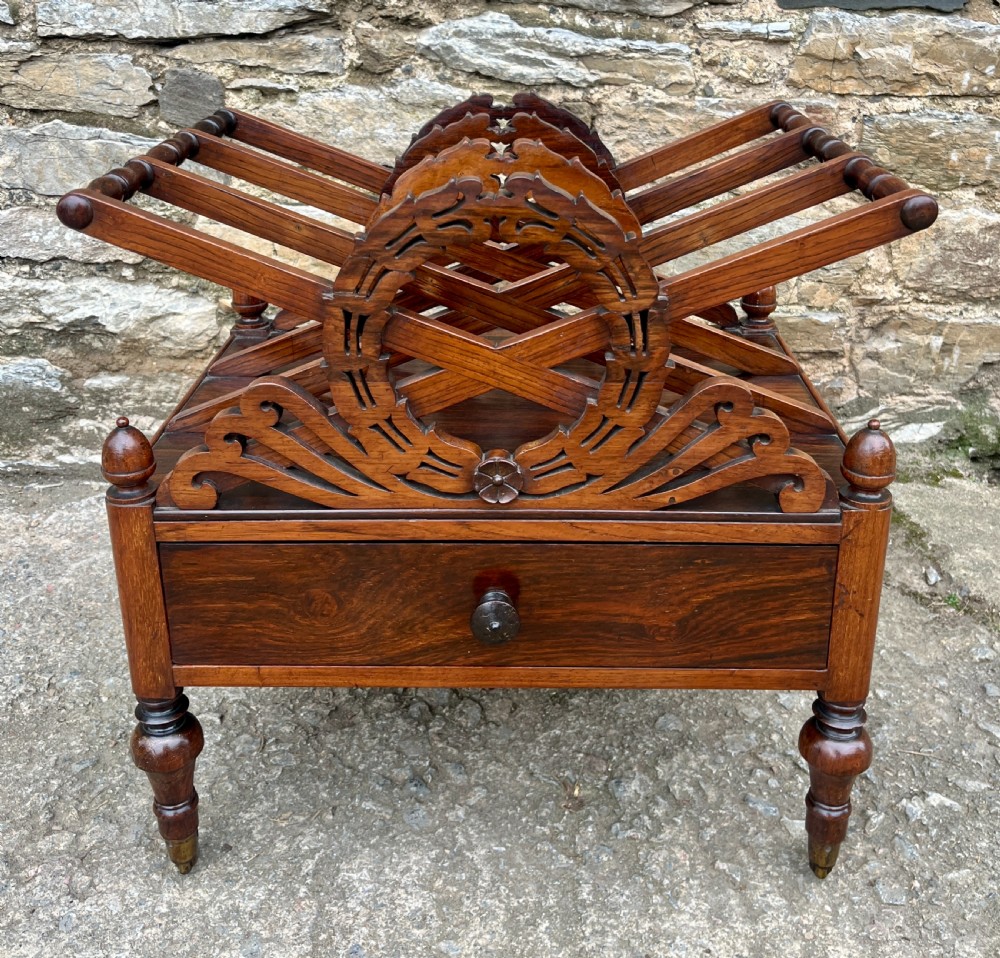 A Regency rosewood music canterbury featuring carved wreath motif side panels, slatted partitions, a single base drawer, and turned legs on brass castors.