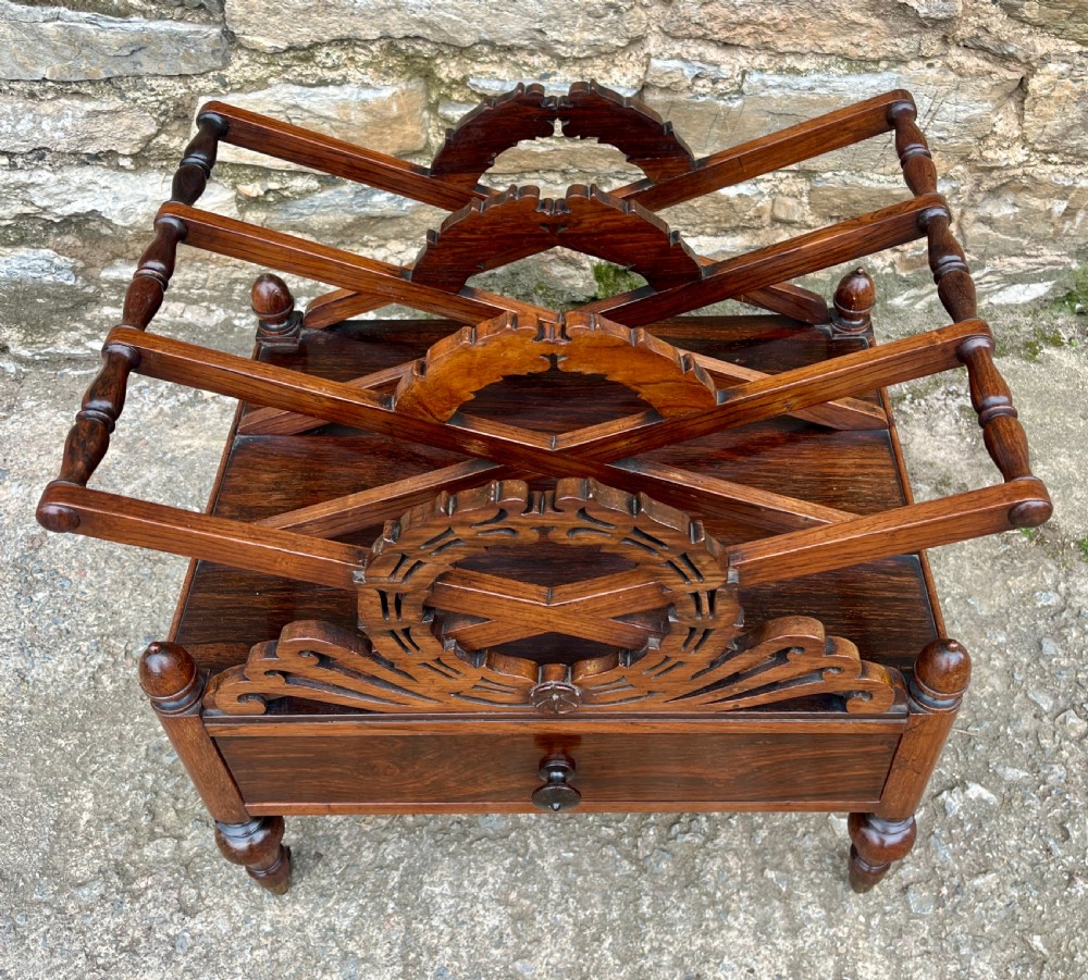 A Regency rosewood music canterbury featuring carved wreath motif side panels, slatted partitions, a single base drawer, and turned legs on brass castors. View II
