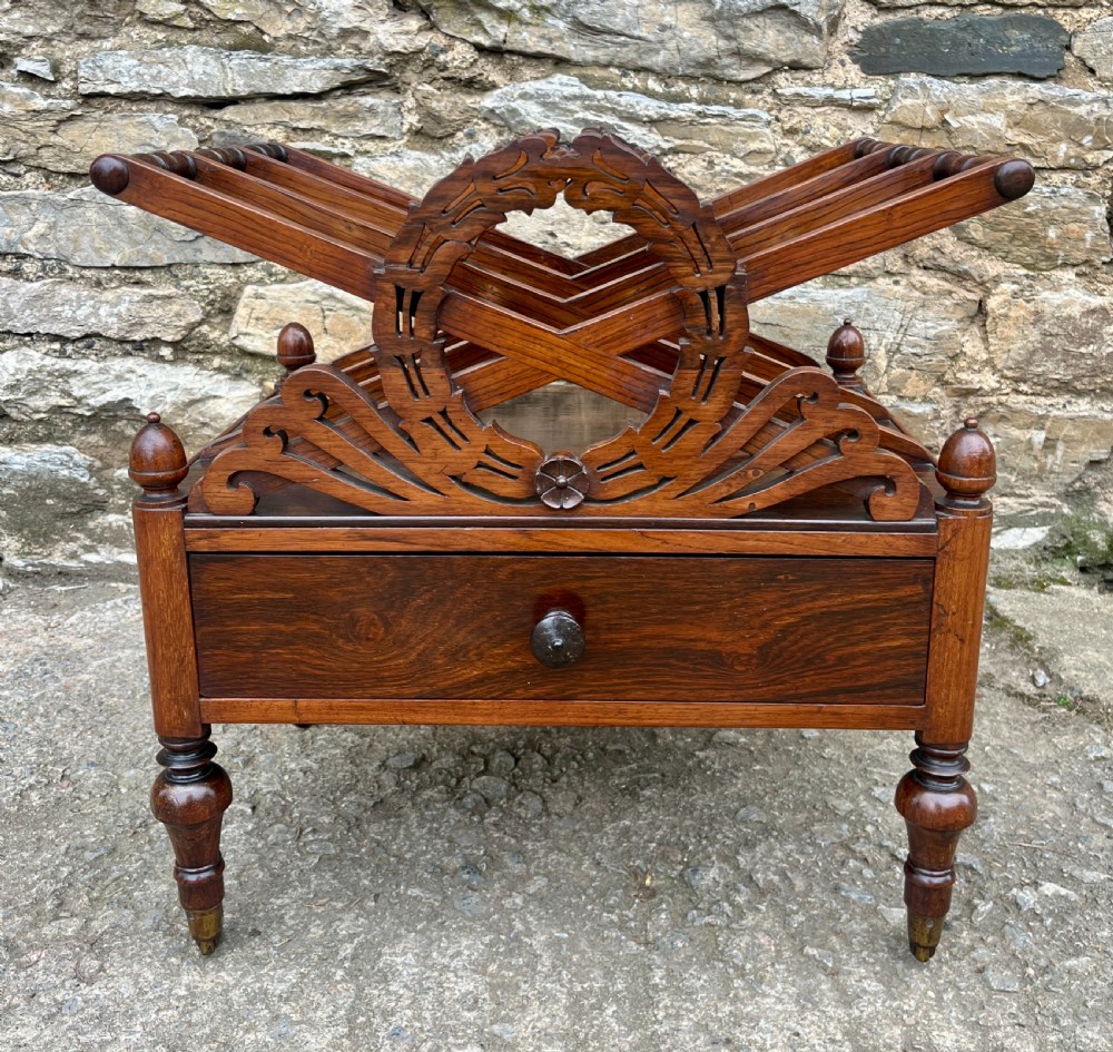 A Regency rosewood music canterbury featuring carved wreath motif side panels, slatted partitions, a single base drawer, and turned legs on brass castors. View III