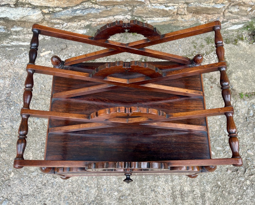A Regency rosewood music canterbury featuring carved wreath motif side panels, slatted partitions, a single base drawer, and turned legs on brass castors. View IV