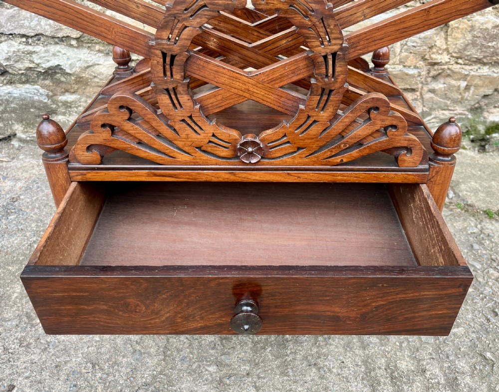 A Regency rosewood music canterbury featuring carved wreath motif side panels, slatted partitions, a single base drawer, and turned legs on brass castors. View V