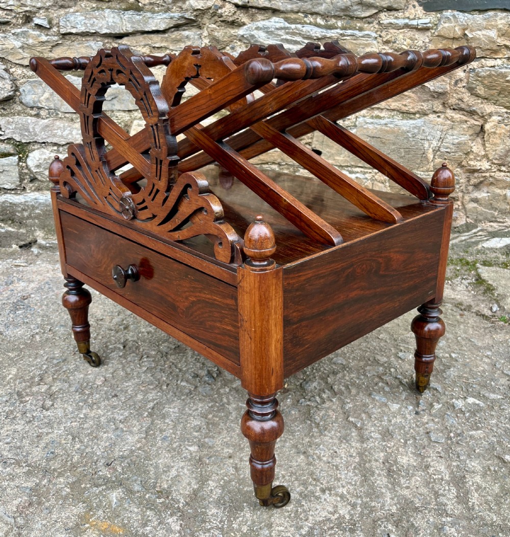 A Regency rosewood music canterbury featuring carved wreath motif side panels, slatted partitions, a single base drawer, and turned legs on brass castors. View VI