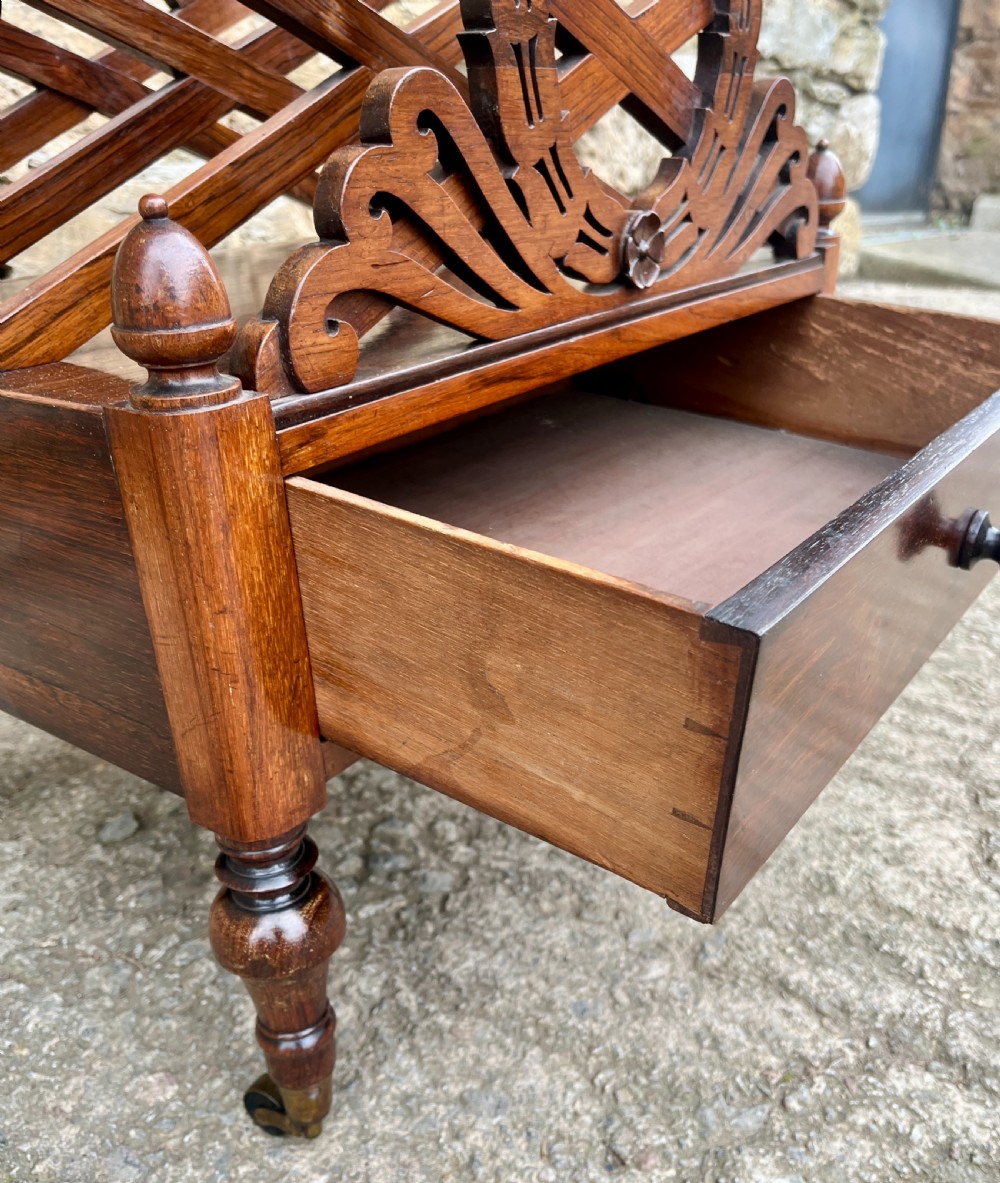 A Regency rosewood music canterbury featuring carved wreath motif side panels, slatted partitions, a single base drawer, and turned legs on brass castors. View VII