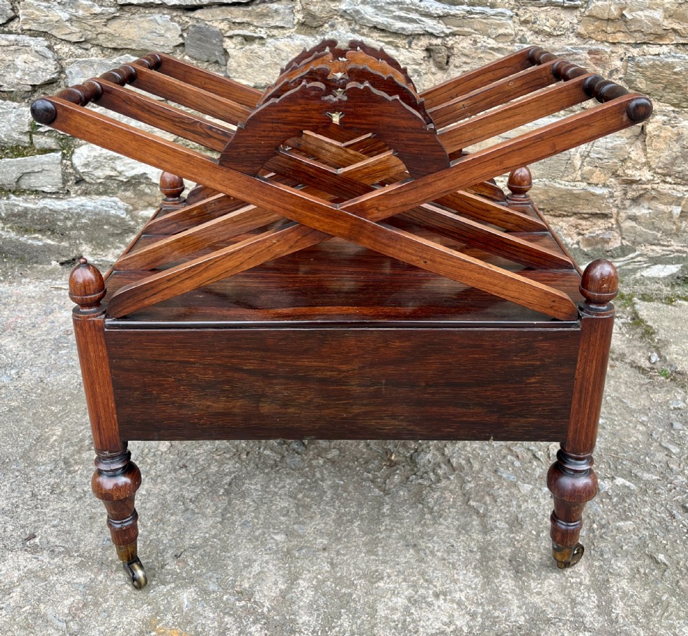 A Regency rosewood music canterbury featuring carved wreath motif side panels, slatted partitions, a single base drawer, and turned legs on brass castors. View IX