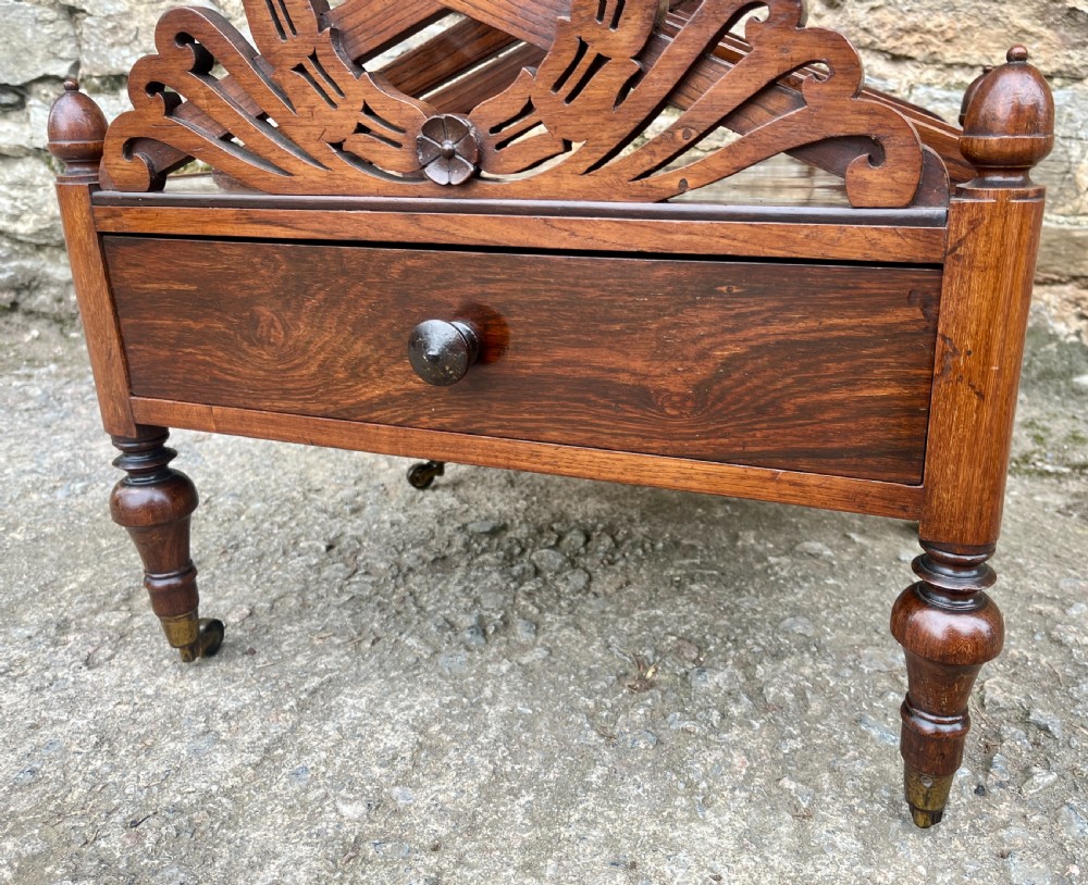 A Regency rosewood music canterbury featuring carved wreath motif side panels, slatted partitions, a single base drawer, and turned legs on brass castors. View X