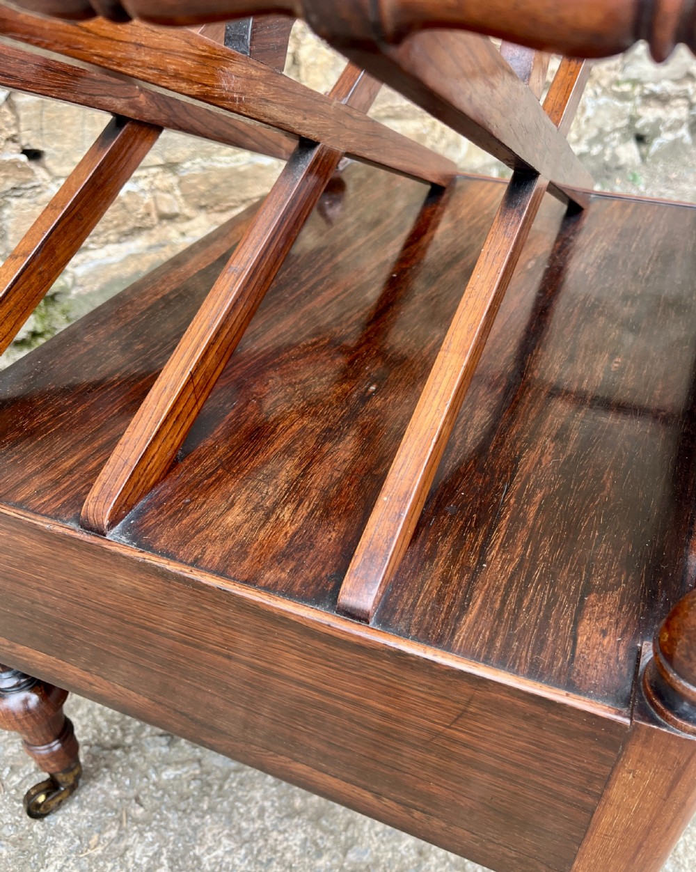 A Regency rosewood music canterbury featuring carved wreath motif side panels, slatted partitions, a single base drawer, and turned legs on brass castors. View XIV