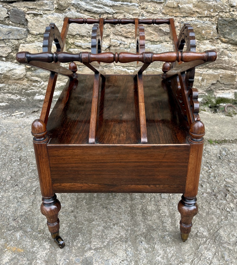 A Regency rosewood music canterbury featuring carved wreath motif side panels, slatted partitions, a single base drawer, and turned legs on brass castors. View XVII