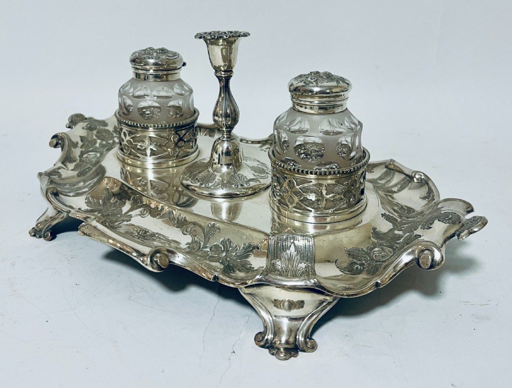 A Victorian silver-plated inkstand featuring an ornate tray with scrolled feet, two cut-glass inkwells in pierced holders, and a central taperstick. View V