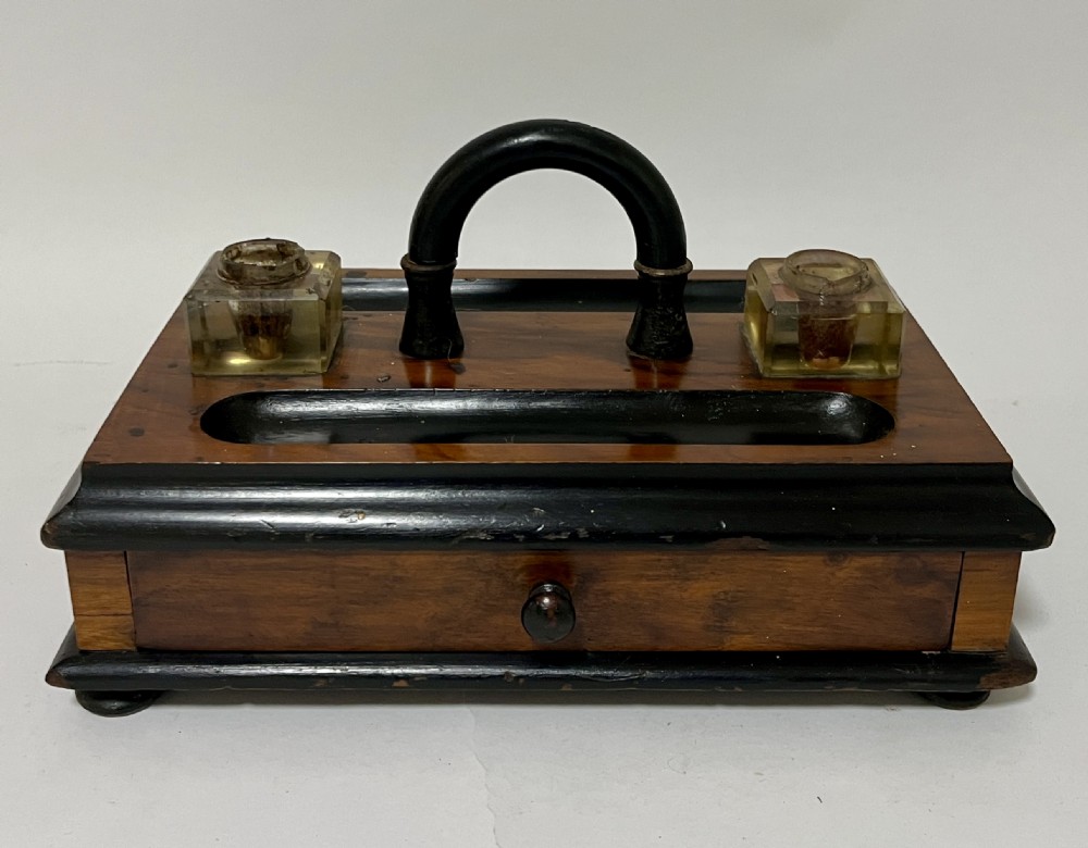 A Victorian wooden desk stand featuring two square glass inkwells, a central black arched handle, an integrated pen tray, and a single lower drawer.