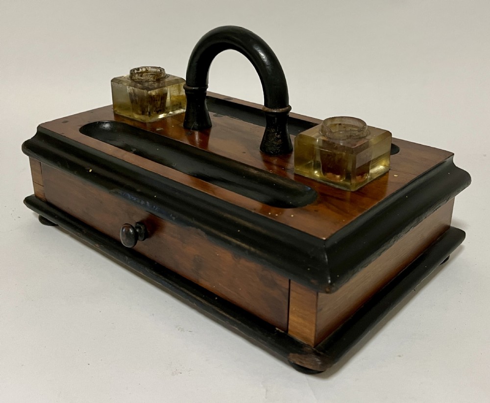 A Victorian wooden desk stand featuring two square glass inkwells, a central black arched handle, an integrated pen tray, and a single lower drawer. View II