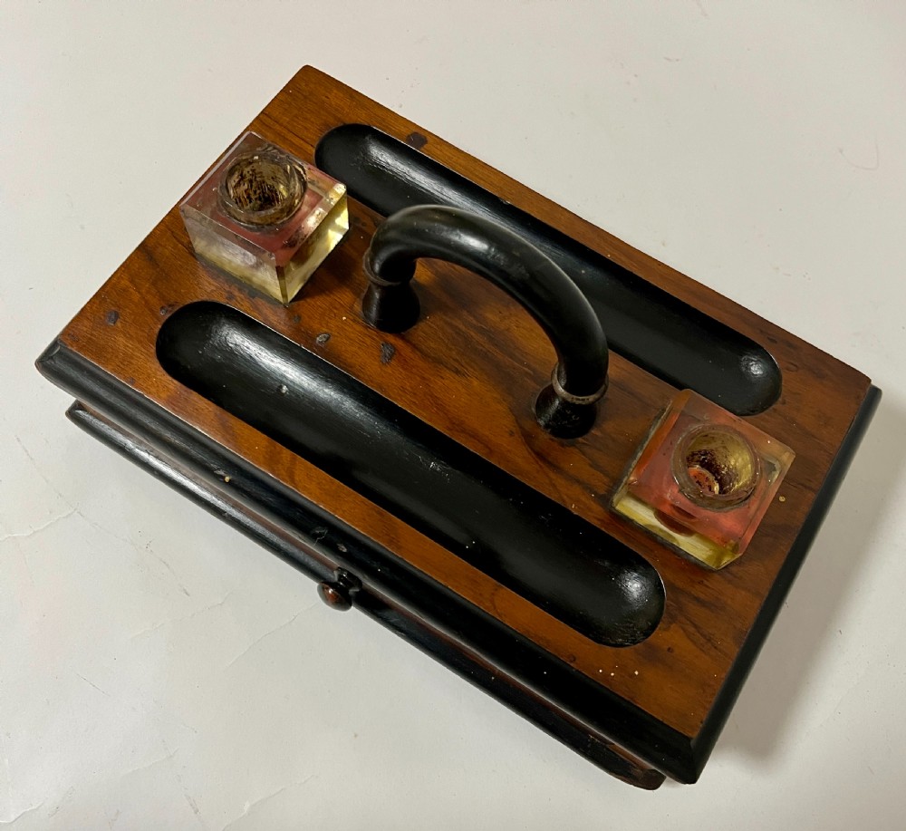 A Victorian wooden desk stand featuring two square glass inkwells, a central black arched handle, an integrated pen tray, and a single lower drawer. View VI