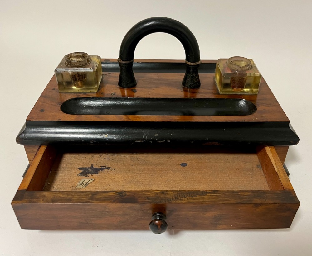 A Victorian wooden desk stand featuring two square glass inkwells, a central black arched handle, an integrated pen tray, and a single lower drawer. View IX