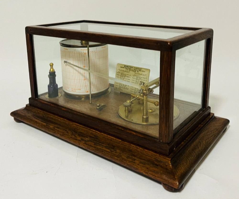 An antique barograph featuring an oak frame with glass side panels enclosing a revolving paper chart drum and polished brass mechanism. View III