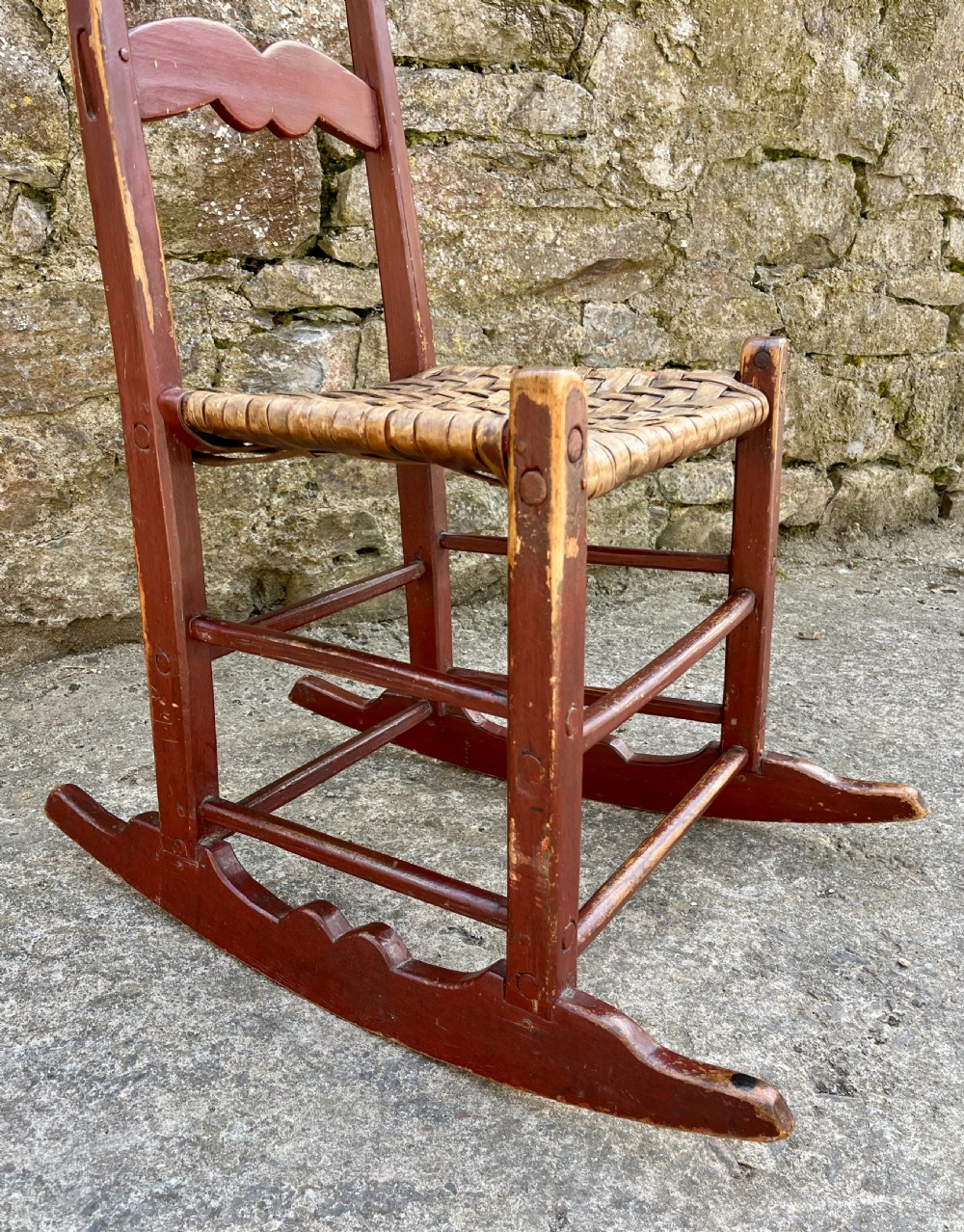 A primitive red-painted wooden rocking chair featuring a woven splint seat and scalloped back slats. View VII
