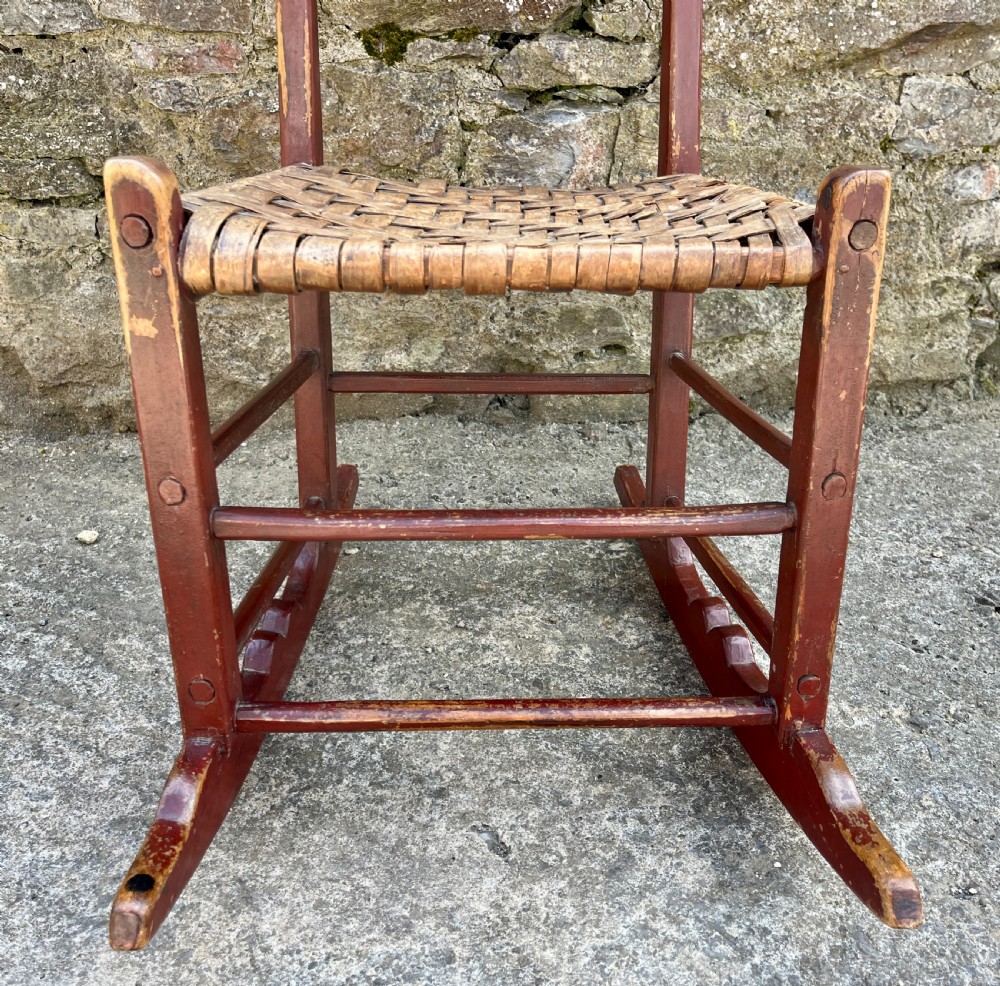 A primitive red-painted wooden rocking chair featuring a woven splint seat and scalloped back slats. View X