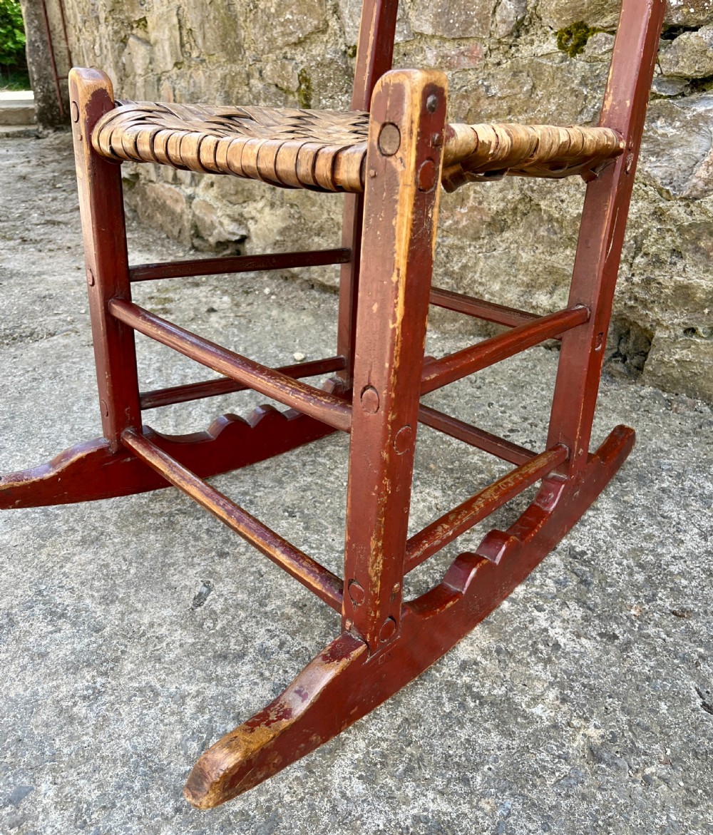A primitive red-painted wooden rocking chair featuring a woven splint seat and scalloped back slats. View XI